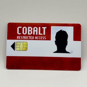 May include: A rectangular card with a red and white design. The word "COBALT" is printed in white block letters above "RESTRICTED ACCESS". A black silhouette of a person is on the right side. A gold-colored chip is on the left.