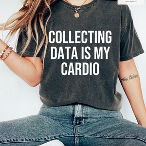 May include: A charcoal grey short-sleeved t-shirt featuring the white text COLLECTING DATA IS MY CARDIO printed on the front. The shirt has a crew neckline and a relaxed fit, paired with blue denim jeans and white sneakers.