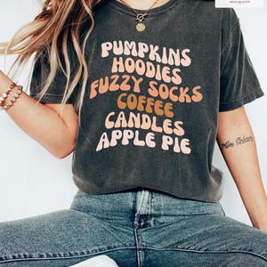 May include: A charcoal grey short-sleeve t-shirt featuring the text PUMPKINS HOODIES FUZZY SOCKS COFFEE CANDLES APPLE PIE in a wavy, retro-style font. The shirt has a crew neckline and a vintage-washed appearance with a soft, worn-in texture.
