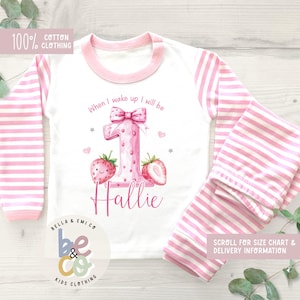 May include: A white and pink striped pajama set for a child. The top has pink sleeves and a pink collar. The front of the top features a large pink number 1 with strawberries and the name "Hallie". The text "When I wake up I will be" is above the number.