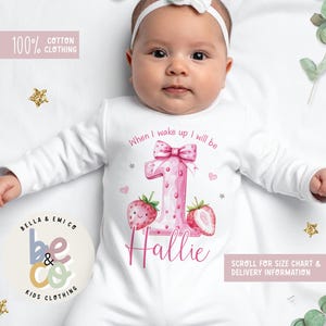 May include: White baby onesie with a pink number one design, strawberries, and the name "Hallie." The onesie has the text "When I wake up I will be." The words "100% Cotton Clothing" and "Bella & Emi Co Kids Clothing" are also visible.