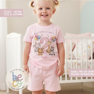 Peut inclure: Un ensemble t-shirt à manches courtes et short rose clair avec un motif représentant un grand chiffre deux, des ballons et des animaux dessinés dont un éléphant, un lion, une girafe et un zèbre. Le texte indique When I wake up I will be et Skyla.