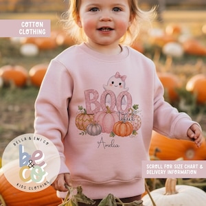 May include: A light pink long-sleeve sweatshirt featuring a graphic of a ghost with a bow, pumpkins, and the text "BOO tiful" along with the name "Amelia" printed in black script. The sweatshirt has a crew neckline and ribbed cuffs.