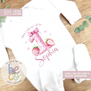 May include: White baby onesie with long sleeves. The onesie features a large pink number one with strawberry accents, the name "Sophia", and the text "When I wake up I will be". The onesie is made of 100% cotton.