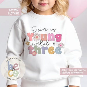May include: White cotton sweatshirt with the text "Erin is Young Wild & Three" in colorful, playful lettering. The design includes floral accents and the brand logo "BELLA & EMICO KIDS CLOTHING". The sweatshirt is part of a children's clothing line.