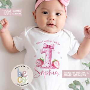May include: A white cotton baby onesie with a pink strawberry-themed design for a first birthday. The design includes a large number 1, strawberries, and the name "Sophia". Text on the onesie reads "When I wake up I will be".