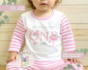 May include: A white and pink striped pyjama set for a one-year-old. The top has the word "ONE" with bows and a crown, and the name "Lottie". The text "100% Cotton Clothing" is visible.