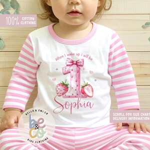 May include: White and pink striped pajama set for a child. The long-sleeved top features a large pink number "1" with a bow, strawberries, and the name "Sophia". The text "When I wake up I will be" is above the number. The set is made of 100% cotton.