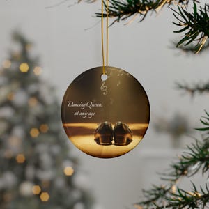 May include: Round Christmas ornament with a gold string, featuring a pair of brown tap shoes on a wooden floor. The text "Dancing Queen, at any age" is displayed in elegant script. Musical notes are also visible.