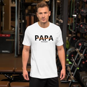 Könnte beinhalten: Weißes T-Shirt mit dem Wort "PAPA" in schwarzen Blockbuchstaben, darunter "Wunschjahr" in kleineren schwarzen Buchstaben. Das Wort "Wunschname" ist in orangefarbener Schrift über "Wunschjahr" geschrieben. Das T-Shirt wird von einer Person in einem Fitnessstudio getragen.