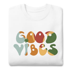 Könnte beinhalten: Weißes Sweatshirt mit dem Schriftzug "GOOD VIBES" in farbenfrohen Buchstaben im Retro-Stil. Die Buchstaben sind in Orange-, Rot-, Grün- und Blautönen gehalten, mit einer verspielten, abgerundeten Schriftart. Ein lustiges und positives Design.