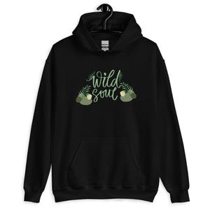 Könnte beinhalten: Schwarzer Kapuzenpullover mit dem Schriftzug "wild soul" in Schreibschrift, verziert mit grünen Farn- und Blattmotiven. Der Pullover hat eine Vordertasche und eine Kapuze mit Kordelzug.