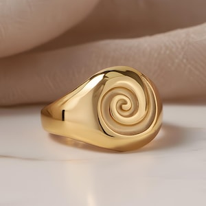 Puede incluir: Un anillo dorado con un diseño en espiral. El anillo tiene una superficie lisa y brillante que refleja la luz. La espiral está centrada en la parte superior del anillo. El fondo es neutro.