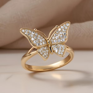 Könnte beinhalten: Ein goldfarbener Ring mit einem Schmetterlingsdesign. Die Schmetterlingsflügel sind mit kleinen, klaren Edelsteinen besetzt. Der Ring wird vor einem neutralen Hintergrund präsentiert, um die Details hervorzuheben.