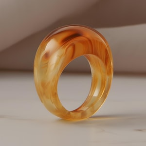 May include: A translucent, amber-colored ring with a rounded, oval shape. The ring has a marbled effect, with swirls of lighter and darker amber tones. The ring is set against a neutral background, highlighting its color and design.