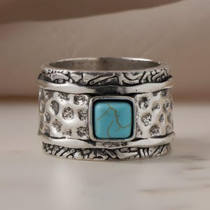 May include: A silver-toned ring with a textured band and a square, turquoise-colored stone. The ring has an antique or vintage style, with detailed patterns around the edges and on the band. The turquoise stone is the focal point.