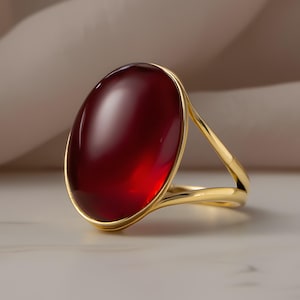 May include: A gold ring featuring a large, oval-shaped, deep red gemstone. The ring's band is a simple, polished gold, and the gemstone is set in a gold bezel. The ring is displayed on a light-colored surface.