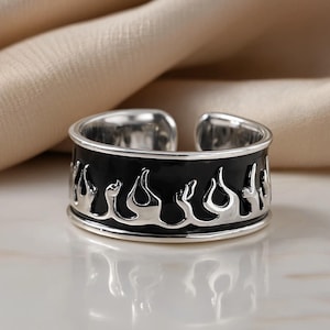Può includere: Un anello aperto color argento caratterizzato da un'ampia superficie piatta con un inserto in smalto nero. L'anello è decorato con un motivo a fiamme in argento lucido in rilievo che avvolge l'intera circonferenza, creando un design ad alto contrasto.