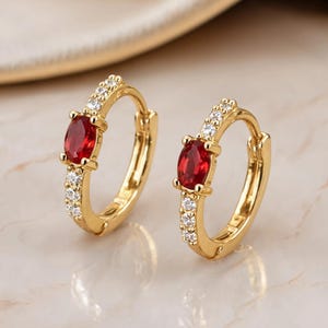 May include: A pair of gold hoop earrings featuring a central oval-cut red gemstone flanked by small clear white stones along the front of the hoop. The earrings have a hinged closure mechanism and a polished metallic finish on the gold-tone metal.