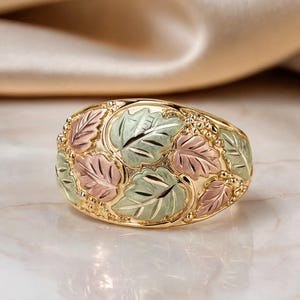 May include: A multi-tone gold ring featuring a detailed cluster of sculpted leaves in alternating shades of yellow, rose, and green gold. The band is wide and domed, showcasing a textured botanical motif with small gold bead accents between the leaves.