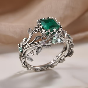 Könnte beinhalten: Ein silberner Ring mit einem smaragdgrünen Edelstein im Smaragdschliff. Der Ring hat aufwendige Blatt- und Rankenmuster, kleine grüne Akzentsteine und ein zartes Band. Das Design ist aufwendig und von der Natur inspiriert.