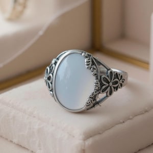 Könnte beinhalten: Ein silberner Ring mit einem ovalen, hellblauen Edelstein. Der Ring hat ein Blumenmuster um den Edelstein und auf dem Band. Der Ring wird auf einer weißen, strukturierten Oberfläche präsentiert.