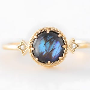 Könnte beinhalten: Ein goldener Ring mit einem runden, irisierenden blauen Edelstein. Der Edelstein ist in einem strukturierten, kreisförmigen Rahmen gefasst. Kleine sternförmige Akzente mit einem einzelnen weißen Stein befinden sich auf beiden Seiten des Edelsteins.