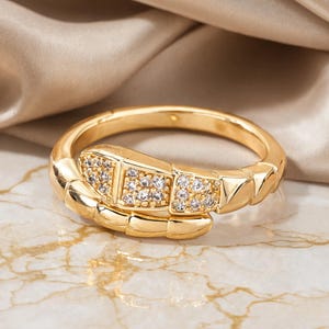 Può includere: Un anello color oro a forma di serpente avvolto con un corpo segmentato. La testa del serpente è decorata con piccole pietre chiare e scintillanti incastonate nella superficie metallica, creando un aspetto strutturato e riflettente sull'anello.