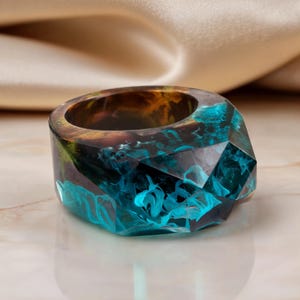 May include: A faceted resin ring featuring a deep amber and brown upper section that transitions into a vibrant, swirling turquoise blue base. The geometric, multi-angled surface creates a crystalline appearance with translucent color depth.