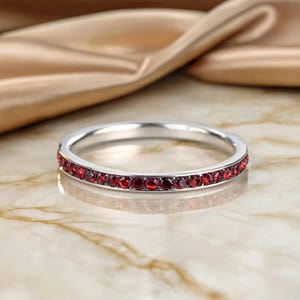 Puede incluir: Un anillo de banda de metal plateado pulido y fino que presenta una fila continua de pequeñas piedras preciosas redondas de color rojo intenso facetadas, engastadas a lo largo de toda la circunferencia exterior de la banda.