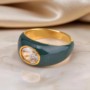Op de afbeelding: Een goudkleurige ring met een brede, donker bosgroene emaille band. Het midden van de ring is gezet met een enkele, heldere, ovaalvormige gefacetteerde edelsteen, omlijst door een dunne goudkleurige metalen rand voor een opvallende uitstraling.