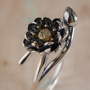 Può includere: Un anello d'argento con un design floreale dettagliato con petali neri e un centro color oro. L'anello include anche un bocciolo, creando un'estetica ispirata alla natura. L'anello è posto su uno sfondo morbido e neutro.