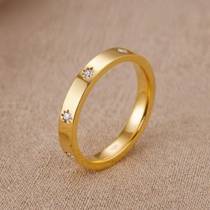 Könnte beinhalten: Ein goldfarbener Ring mit flachem Band, verziert mit kleinen, sternförmigen Diamant-Akzenten. Der Ring hat eine polierte Oberfläche. Der Schmuck ist vor einem neutralen Hintergrund platziert.