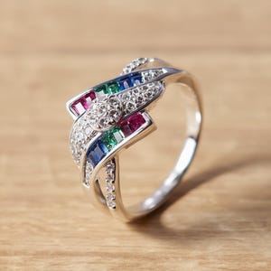 Könnte beinhalten: Silberner Ring mit einzigartigem Design. Er hat eine zentrale Ansammlung kleiner, klarer Steine, umgeben von roten, grünen und blauen rechteckigen Edelsteinen. Der Ring ist zusätzlich mit klaren Steinen besetzt.