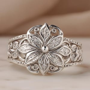 Peut inclure: Une bague en argent ornée d'un motif floral détaillé. La bague présente une grande fleur ouverte avec des détails complexes sur les pétales. La bande est ornée de volutes et de motifs floraux délicats, créant une esthétique vintage.