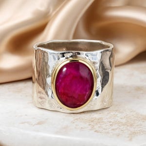 Op de afbeelding: Een brede zilveren ring met een gehamerd, getextureerd oppervlak en een centrale ovaalvormige, donkerroze edelsteen. De edelsteen is gevat in een gladde, gepolijste goudkleurige metalen zetting die contrasteert met de zilveren band.