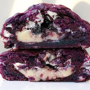 May include: Two halves of a purple cookie, stacked. The cookie has a dark purple exterior and a creamy white filling with dark purple and black inclusions. The cookie is cut to reveal the inside texture and is a sweet treat.