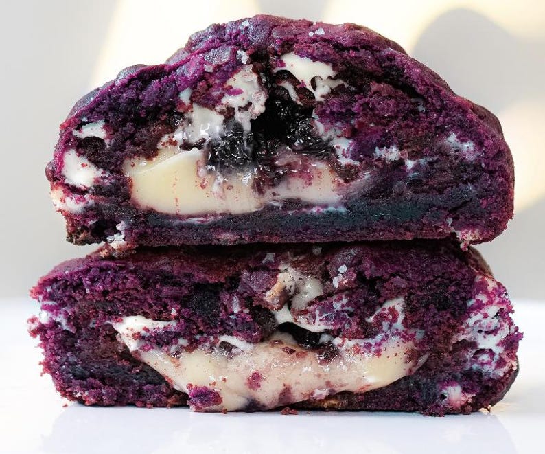 May include: Two halves of a purple cookie stacked on top of each other. The cookie has a dark purple exterior and a creamy white filling with dark purple and black inclusions. The cookie is cut to reveal the inside texture.