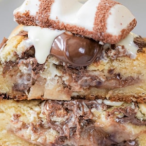 May include: A stack of two halves of a cookie, filled with chocolate and topped with a white chocolate candy bar. The cookie has a golden-brown exterior and a soft, chocolate-filled interior. White chocolate drips down the side.