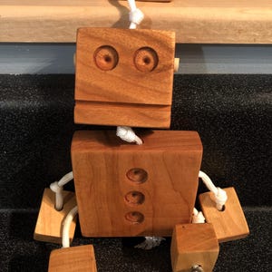 May include: A wooden robot toy with a rectangular head and body, connected by a white rope. The head features two circular eye indentations. The body has three circular indentations. The arms and legs are made of wooden blocks connected by white rope.