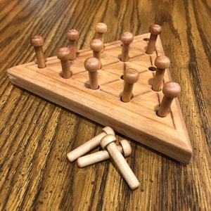 May include: A wooden peg solitaire game with a triangular board and wooden pegs. The board has holes arranged in a grid pattern, with pegs inserted in most of the holes. Three pegs are lying next to the board. The game is set on a wooden table.