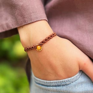 Può includere: Un braccialetto in macramè marrone con un ciondolo in pietra preziosa gialla. Il braccialetto è indossato su un polso, con una persona che indossa una camicia malva e jeans blu. È un gioiello fatto a mano.