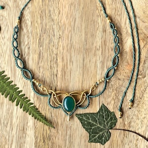 Può includere: Una collana macramè fatta a mano con una pietra verde ovale centrale e dettagli in metallo color oro. La collana presenta un cordino verde scuro con nodi intricati e lunghezza regolabile. Elementi decorativi: un ciondolo a forma di sole dorato e foglie verdi.