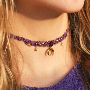 Könnte beinhalten: Ein lila Makramee-Choker-Halskette mit goldenen Akzenten und einem Halbmond-Anhänger. Die Halskette hat einen zentralen dunkelvioletten Stein und kleine goldene Tropfenanhänger. Das Design ist aufwendig und im Bohème-Stil gehalten.