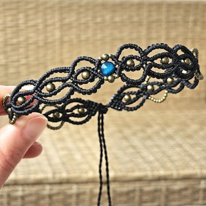 May include: Black macrame bracelet with a central blue bead and small brass beads. The intricate knotwork design creates a delicate, open pattern. The bracelet is held by a hand, showcasing its adjustable tie closure.