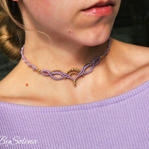 Op de afbeelding: Een delicate lavendelkleurige macramé choker ketting met goudkleurige accenten. De ketting heeft ingewikkelde knopen en een centraal decoratief element. De afbeelding toont de ketting op een persoon die een lavendelkleurige top draagt. De tekst "By Solena" is zichtbaar.