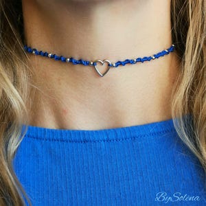 Könnte beinhalten: Ein blauer Makramee-Choker mit einem silbernen Herzanhänger und kleinen silbernen Perlen. Die Halskette wird von einer Person mit blonden Haaren und einem blauen Oberteil getragen. Der Etsy-Shop-Name "By Solena" ist sichtbar.