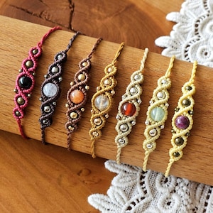 Op de afbeelding: Een verzameling van zeven macramé armbanden in verschillende kleuren, elk met een centrale edelsteen. De armbanden worden gepresenteerd op een houten oppervlak, waarbij de ingewikkelde knoop- en kralenwerk worden getoond. Edelstenen zijn onder andere zwart, wit, oranje, rood, groen en paars.