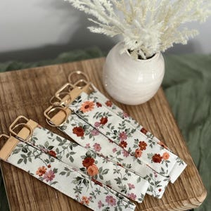 Puede incluir: Llaveros florales con herrajes dorados y detalles de cuero. La tela presenta un fondo blanco con un estampado de flores rojas, rosas y naranjas con hojas verdes. Se muestran sobre una tabla de madera.