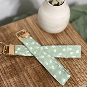 Puede incluir: Dos llaveros con lunares blancos sobre fondo verde claro. Los llaveros tienen herrajes dorados y están sobre una tabla de madera. Un jarrón blanco con flores secas está en el fondo. Miden aproximadamente 15 cm de largo.